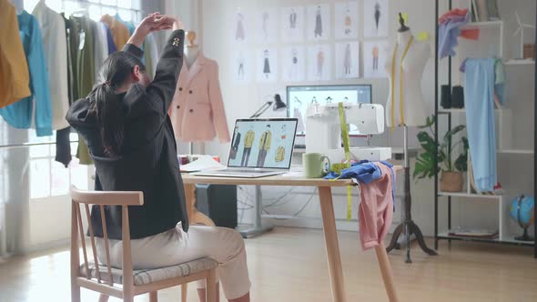 Back View Of Asian Female Designer In Business Suit Stretching While Designing Clothes On A Laptop