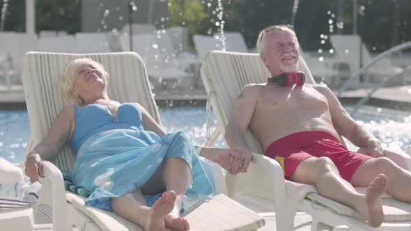 Elderly Positive Couple Lying on Sunbeds Near the Pool Holding Hands and Smiling