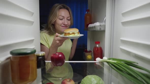 Young Woman Opens the Refrigerator Door at Night and Happy Takes a Burger Instead of an Apple
