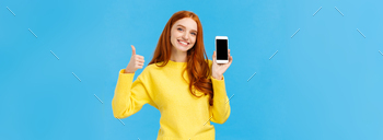 ful application. Cute redhead woman in yellow sweater, like winter holiday sale in online store, showing smartphone display, blue background.