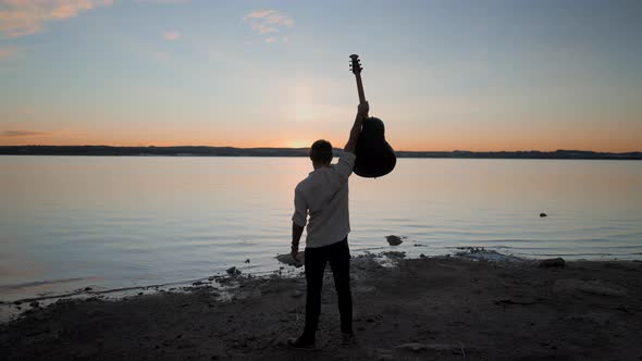 Silhouetted Man Holding a Guitar Raised Up in the Air While Watching Stunning Golden Sunset Over