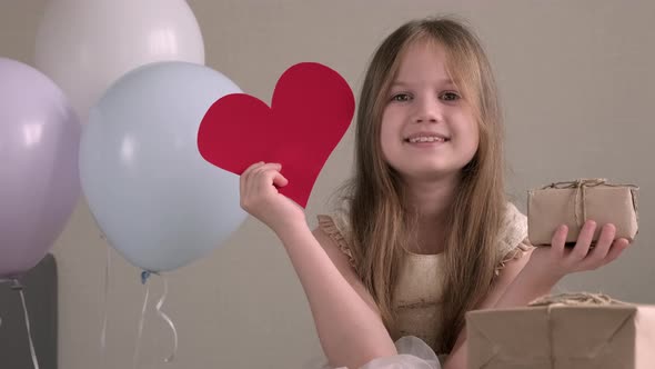 Cute little girl holding big red paper heart and zero waste gift on background of air balloons