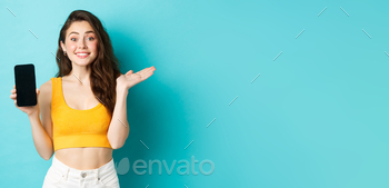 howing emtpy smartphone screen and smiling at camera, standing against blue background.