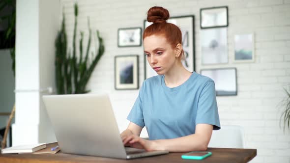 Beautiful Young Redhead Woman Is Working on Laptop at the Desk in Room at Home.