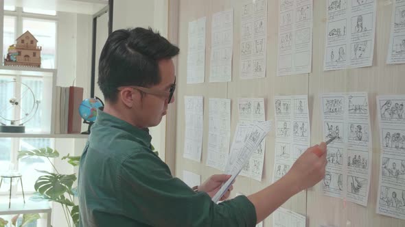 Young Creative Asian Designer Looks At The Storyboard Sketches Covering His Wall