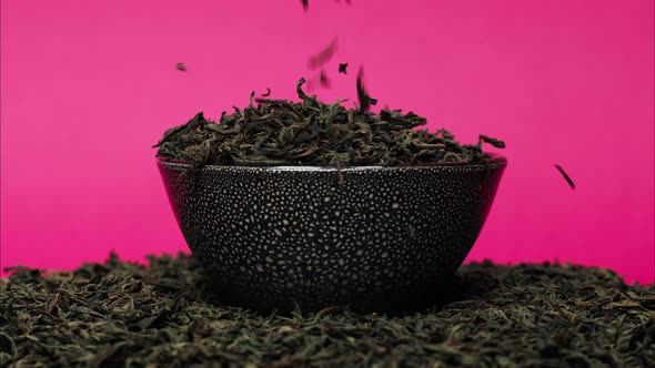 Closeup of Falling Down Dried Tea Leaves Into Black Bowl on Pink Background