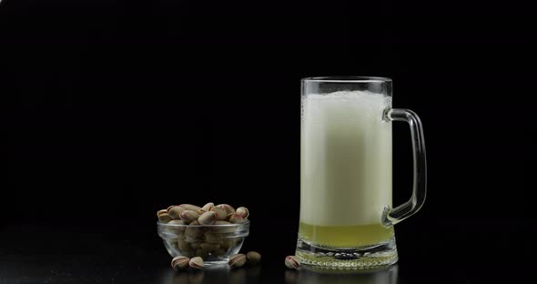 Beer Is Pouring Into Glass on Black Background. Bowl of Pistachios Nuts