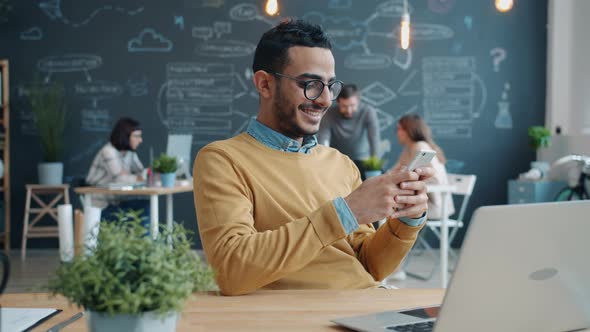 Happy Arabian Guy Using Smartphone and Smiling at Desk in Shared Workplace