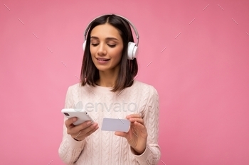 ing pink casual sweater isolated over pink background wall wearing white bluetooth wireless headphones and listening to music and using mobile phone making payment online through credit card looking down.