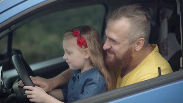 Side View of Happy Little Girl Sitting with Bearded Laughing Man in Car Holding Steering Wheel