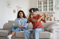 Enthusiastic positive teenage girl with VR glasses with virtual baseball bat sits on couch near mom - PhotoDune Item for Sale