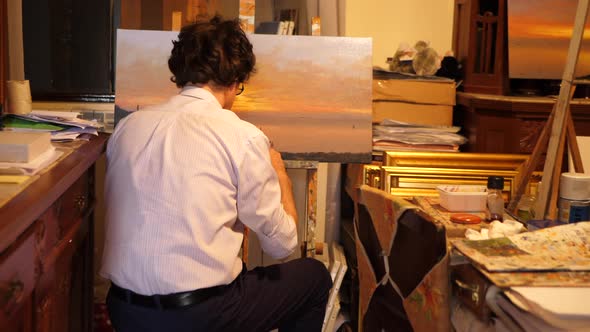 An artist paints an oil painting on canvas inside his art studio.