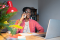 Smiling African guy talking on phone while sitting at desk with laptop, distracted from online study - PhotoDune Item for Sale