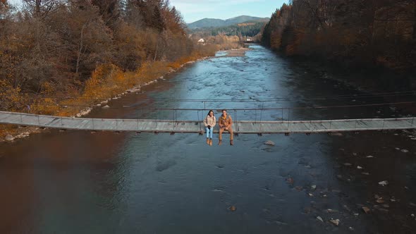 Drone Selfie Footage By Couple Happy Hikers Sitting on Wooden Suspension Bridge Over Mountain River