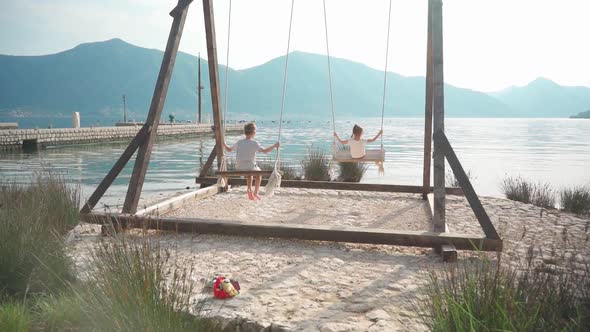 A Girl and a Boy Swing on a Swing Near the Sea with a Beautiful View of the Bay and Mountains