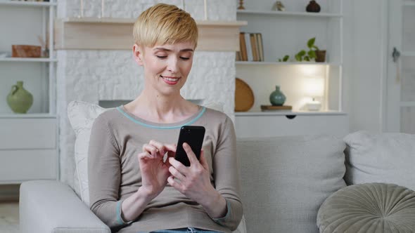 Happy Relaxed Woman Sitting on Sofa Holding Smartphone Looking at Cellphone Screen Surfing Social