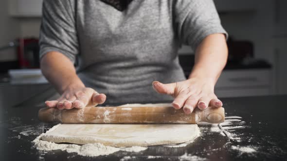 Woman rolling out dough in flour on a black counter top. Lady in gray sweater making pastry dough in