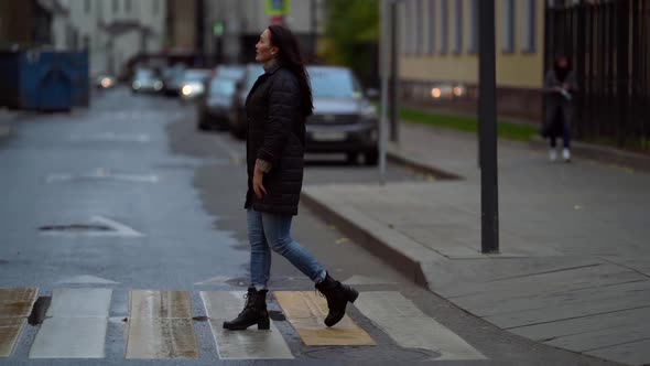 A Middle-aged, Dark-haired Woman in a Black Jacket Walks Along a Pedestrian Crosswalk on a City
