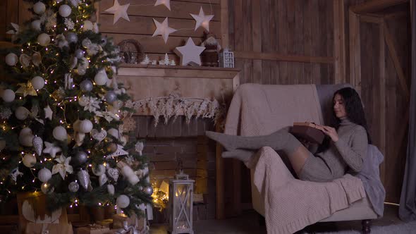 A Girl with Long Hair Opens a Box with a Gift and Rejoices Sitting in a Chair Near the Christmas