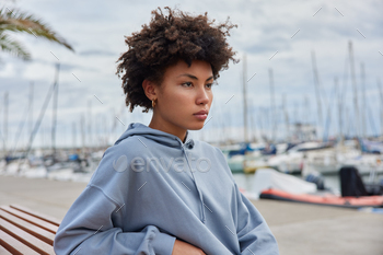 oman dressed in hoodie poses on bench at pier bing deep in thoughts while admiring sea view in harbor looks seriously forward. Outdoor shot.