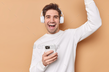  favorite playlist listens music via headphones holds mobile phone feels joyful isolated over beige background boosts mood with favorite song