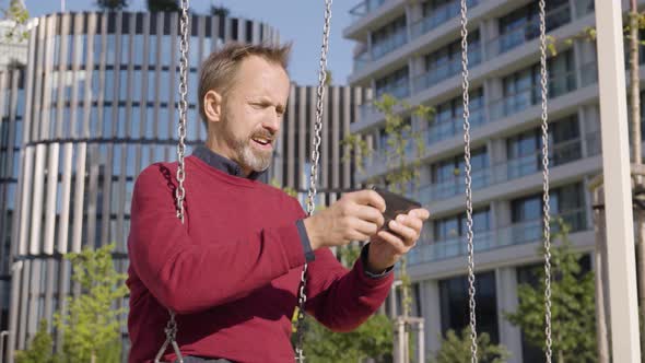 A Middleaged Handsome Caucasian Man Plays a Game on a Smartphone As He Sits on a Swing