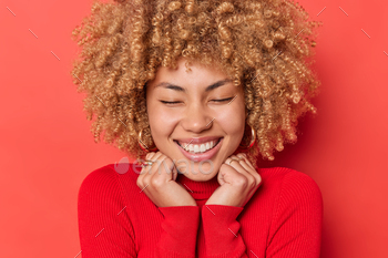 Close up portrait of cheerful curly haired young woman keeps hands on collat of turtleneck closes ey