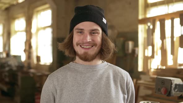 Young Caucasian Male with Red Beard Wearing Black Sock Cap Standing in Joinery and Smiling in