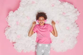 s calm music via headphones dressed in pajama applies beauty patches under eyes poses on white fluffy cloud with candies around. Rest concept