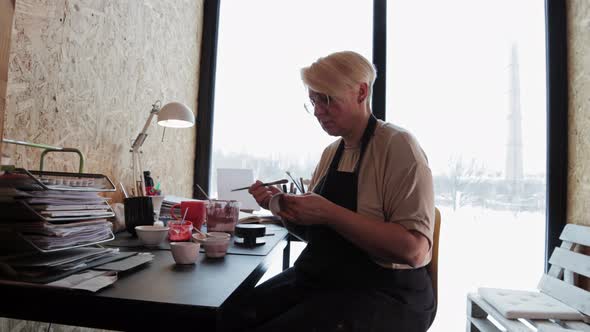 An Elderly Woman Painting Ceramic Mug with a Brush