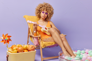 chair plays guitatr to entertain herself wears swimsuit looks sadly uses portable fridge keeps legs in inflated swimming pool isolated on purple wall