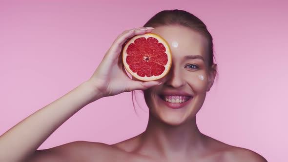 Beautiful woman covers one eye with round slice of orange and smiles, drops of cosmetic cream