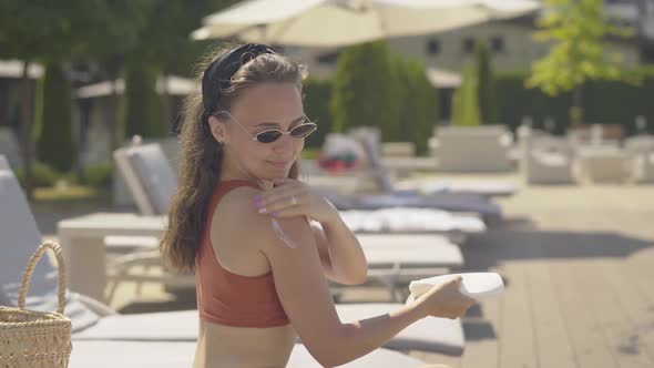 Side View of Confident Positive Girl Applying Suntan Cream on Shoulder and Smiling. Portrait of