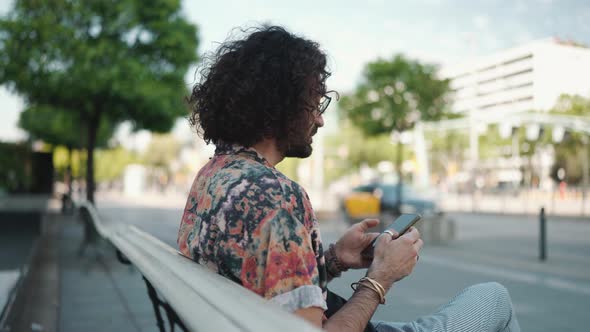 Curly-haired bearded man in eyeglasses texting by mobile