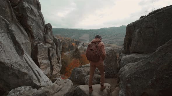 Rear View Man Hiker in Casual Brown Wear with Backpack Walks to Edge of Cliff Enjoys a Beautiful