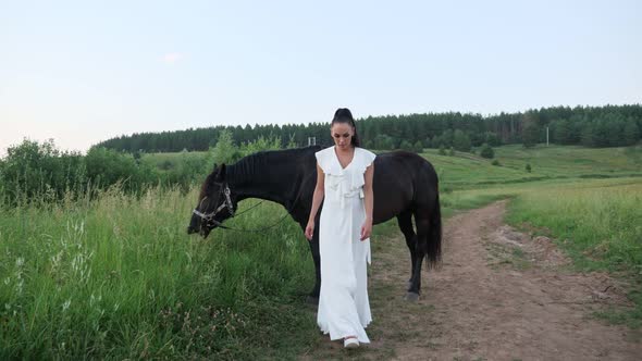 Young Woman Brunette Walks and Black Stallion Eats Grass