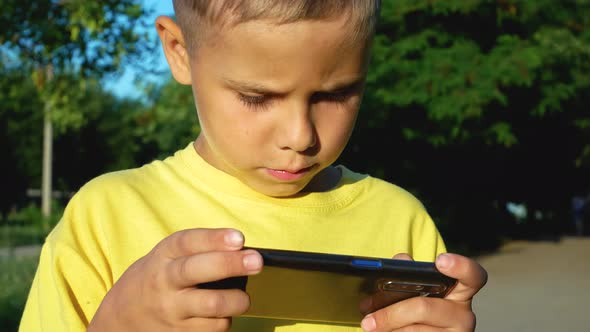 A Cute Boy of 8 Years Old Enthusiastically Plays a Game on His Phone on a Summer Day on the Street