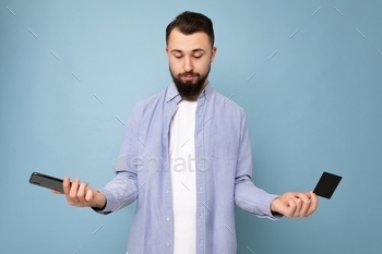 oung man wearing casual blue shirt and white t-shirt isolated over blue background wall holding credit card and using mobile phone making payment looking down and having doubts.