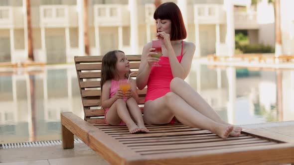 Mom with Cocktail Glass and Her Daughter with Orange Juice Lying on a Wooden Deck Chair