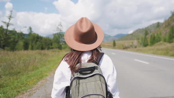 Attractive Brunette Girl in a Hat with a Backpack Travels in the Mountains Hitchhiking, Votes the