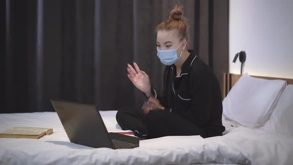 Teenage Caucasian Girl in Covid-19 Face Mask and Pajamas Sitting on Bed and Talking at Laptop