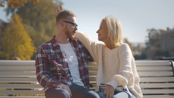 Senior Woman and Adult Son Talking and Hugging Sitting Together on Bench in Park