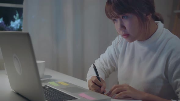 Young asian woman working late using laptop on desk writing notebook document finance and calculator