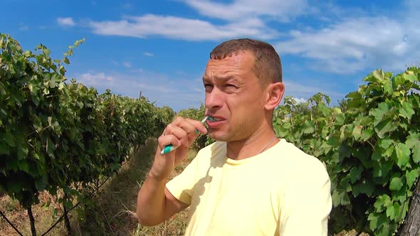 Young man brushes teeth while standing outdoors against backdrop of vineyards.