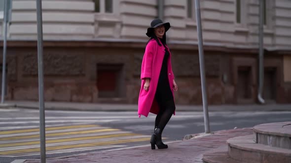 A Beautiful Brunette Woman in a Pink Coat and Black Hat Walks on a Blurry Background of a City