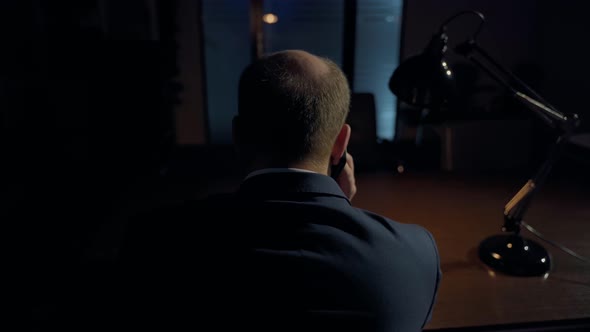 Unrecognizable adult man boss talking phone sitting at table. Office interior and desk lamp visible