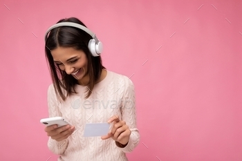 aring pink casual sweater isolated over pink background wall wearing white bluetooth wireless headphones and listening to music and using mobile phone making payment online through credit card looking down. empty space