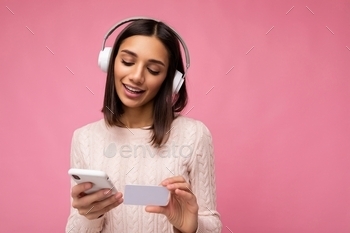 earing pink casual sweater isolated over pink background wall wearing white bluetooth wireless headphones and listening to music and using mobile phone making payment online through credit card looking down.