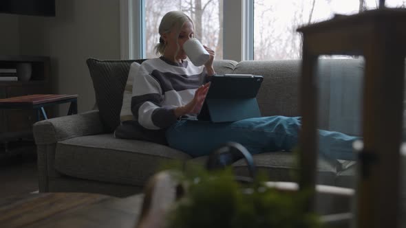 Woman sitting on the couch in front of a bay window, drinking coffee while scrolling on a tablet wit