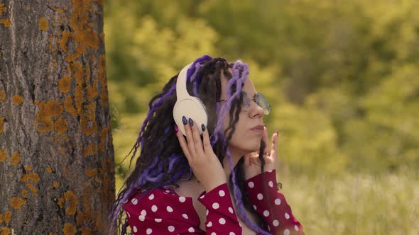 Young Relaxed Woman in Glasses Headphones Sitting Near Tree Listening to Music Dancing Outdoors in